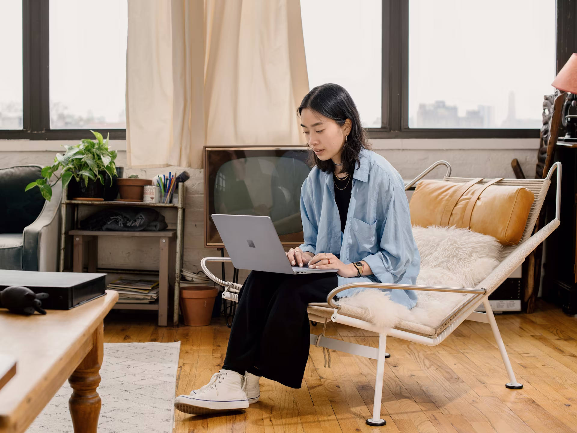 A portrait-style image of a young woman with long, dark hair working at a desk. A laptop, a cup of coffee, and a red book are also visible on the wooden table
