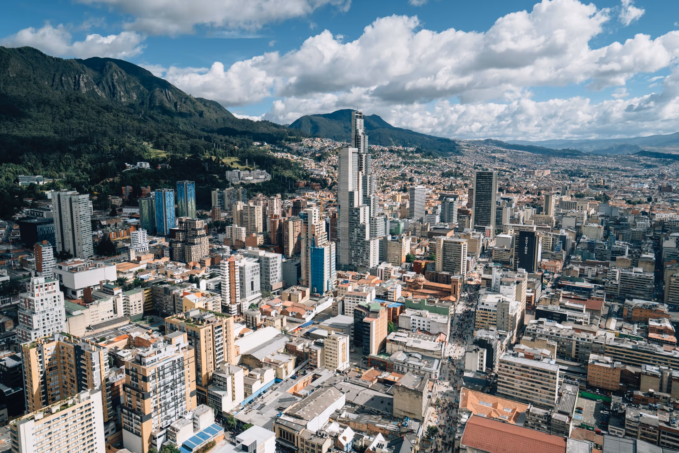 An aerial, wide-angle view of a densely packed, sprawling city at the base of large, dark green, forested mountains. The city features a mix of high-rise and mid-rise commercial and residential buildings, with one distinctive tall, modern skyscraper towering above the rest. The sky is bright with scattered white cumulus clouds.