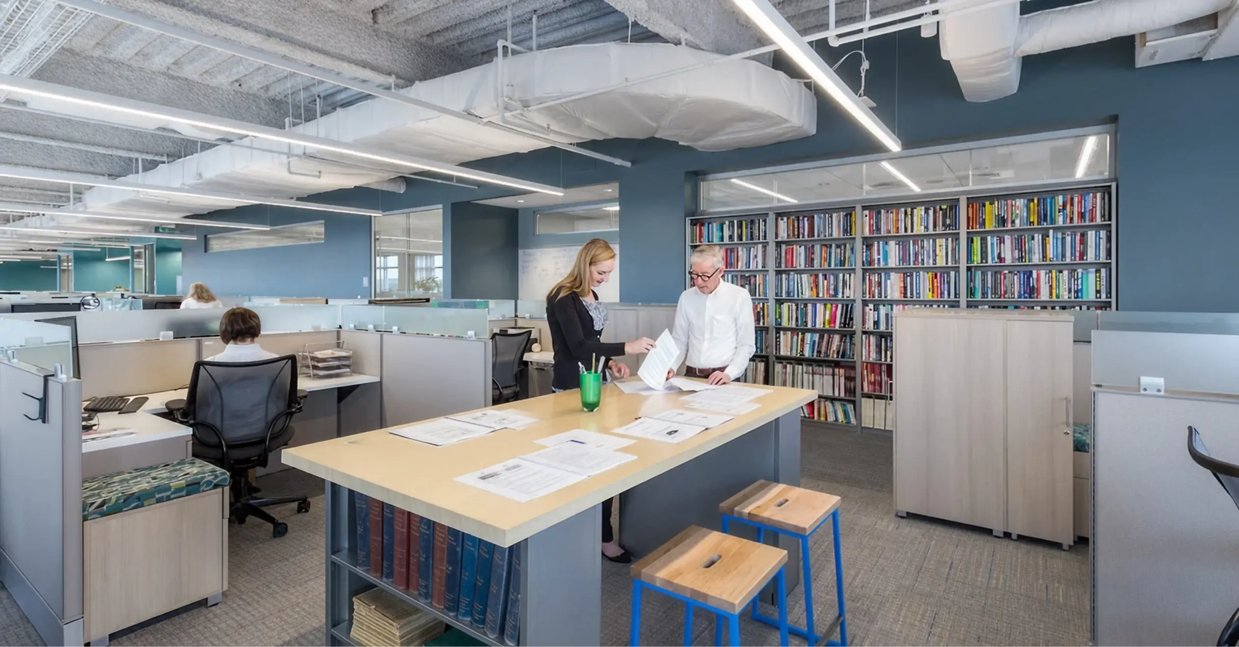 A bright, wide shot of a modern open-plan office with exposed ceilings, long linear lighting fixtures, and light gray cubicle walls. The office features blue-painted accent walls. In the foreground, a woman in a black and white top is standing with an older man in a white collared shirt, reviewing papers spread across a central communal table. The table has a bookshelf base and is accompanied by two stools with blue frames. Along the back wall is a large, built-in bookshelf filled with colorful books.