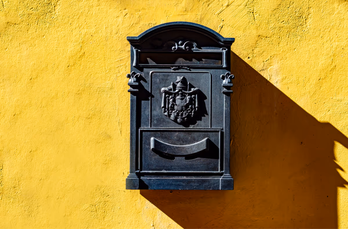 A dramatic, high-contrast photograph of an ornate, black metal mailbox mounted on a brightly textured yellow stucco wall. The mailbox, which features a coat of arms or crest, is strongly lit from the side, casting a sharp, elongated shadow on the vibrant yellow background.
