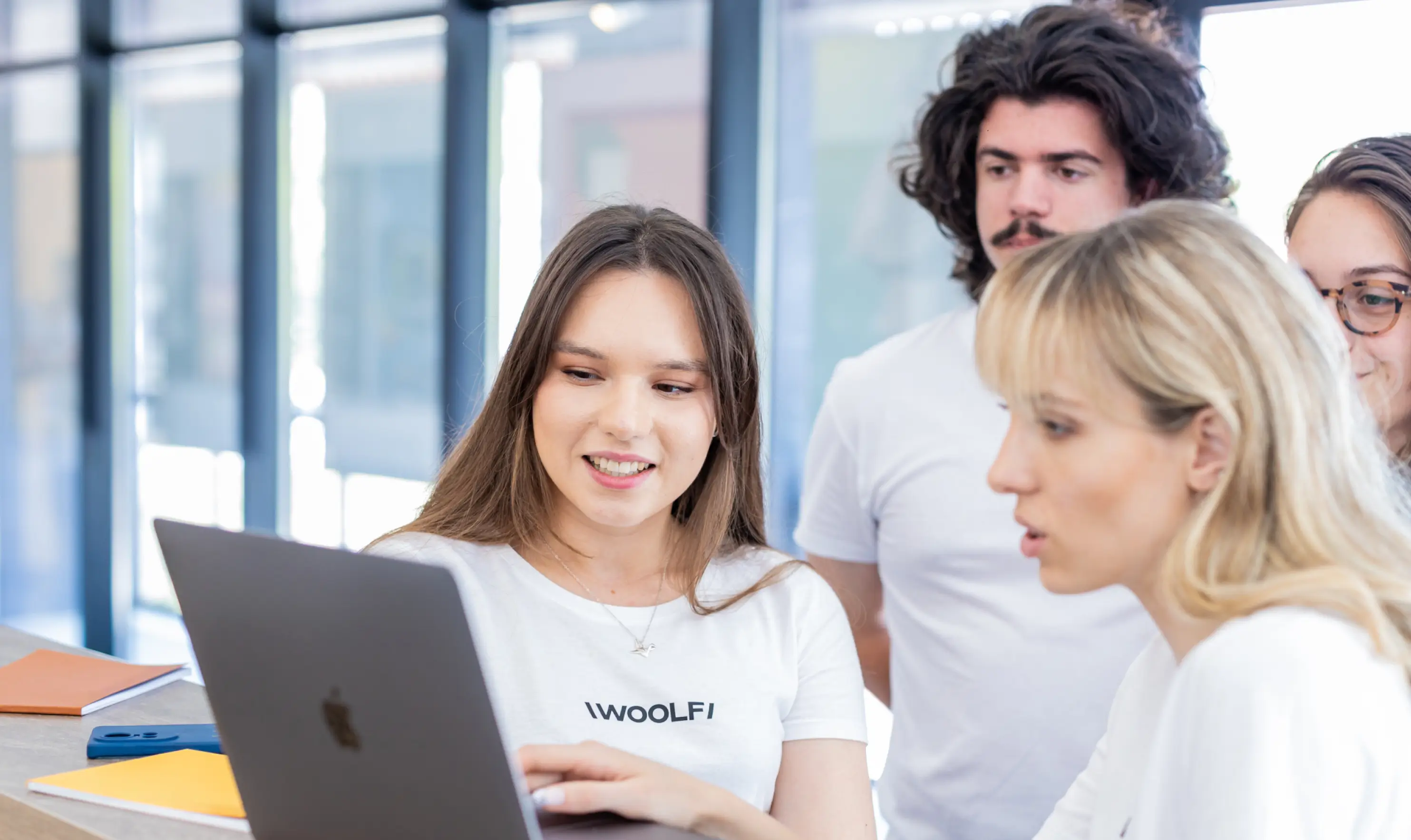 A candid photograph of four young adults collaborating around a MacBook laptop on a tabletop in a modern office or study space. A smiling woman in the foreground, wearing a white t-shirt with the word "WOOLF" printed on it, operates the laptop. Another woman looks at the screen from the right, and two individuals (a man with dark, curly hair and mustache, and a third person) stand behind them, observing the screen. The background is brightly lit with large windows or glass partitions.