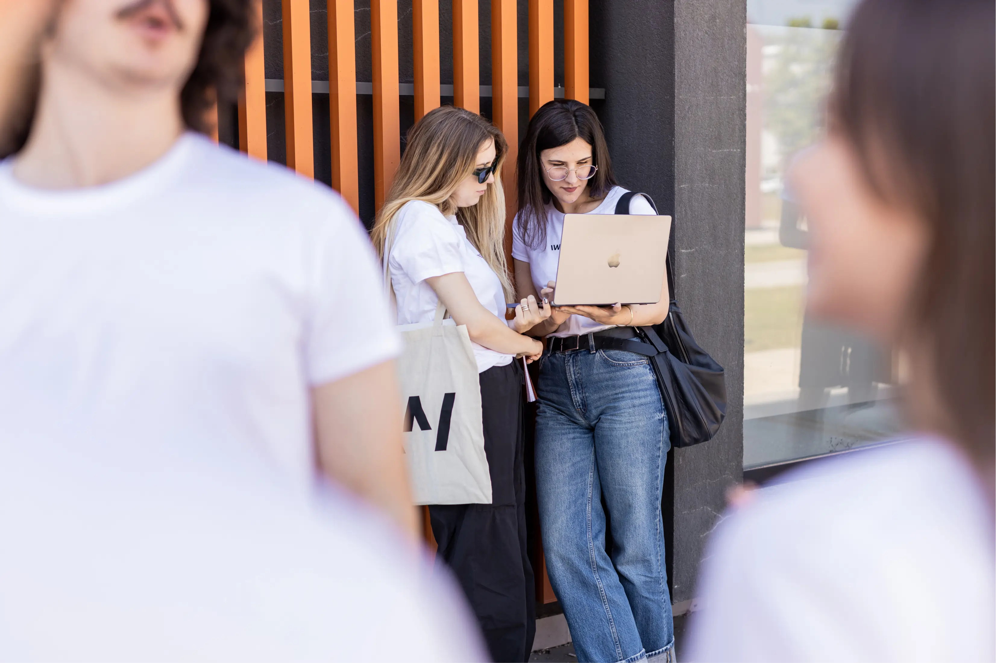 A candid, outdoor photograph focusing on two young women looking at a laptop. The woman on the right, wearing a white t-shirt, jeans, and glasses, holds a gold-colored laptop. The woman on the left, also in a white t-shirt, leans in to look at the screen while holding a large tote bag. They are standing next to a wall with orange vertical slat detailing. The foreground is blurred by the out-of-focus backs of two other people wearing white t-shirts.