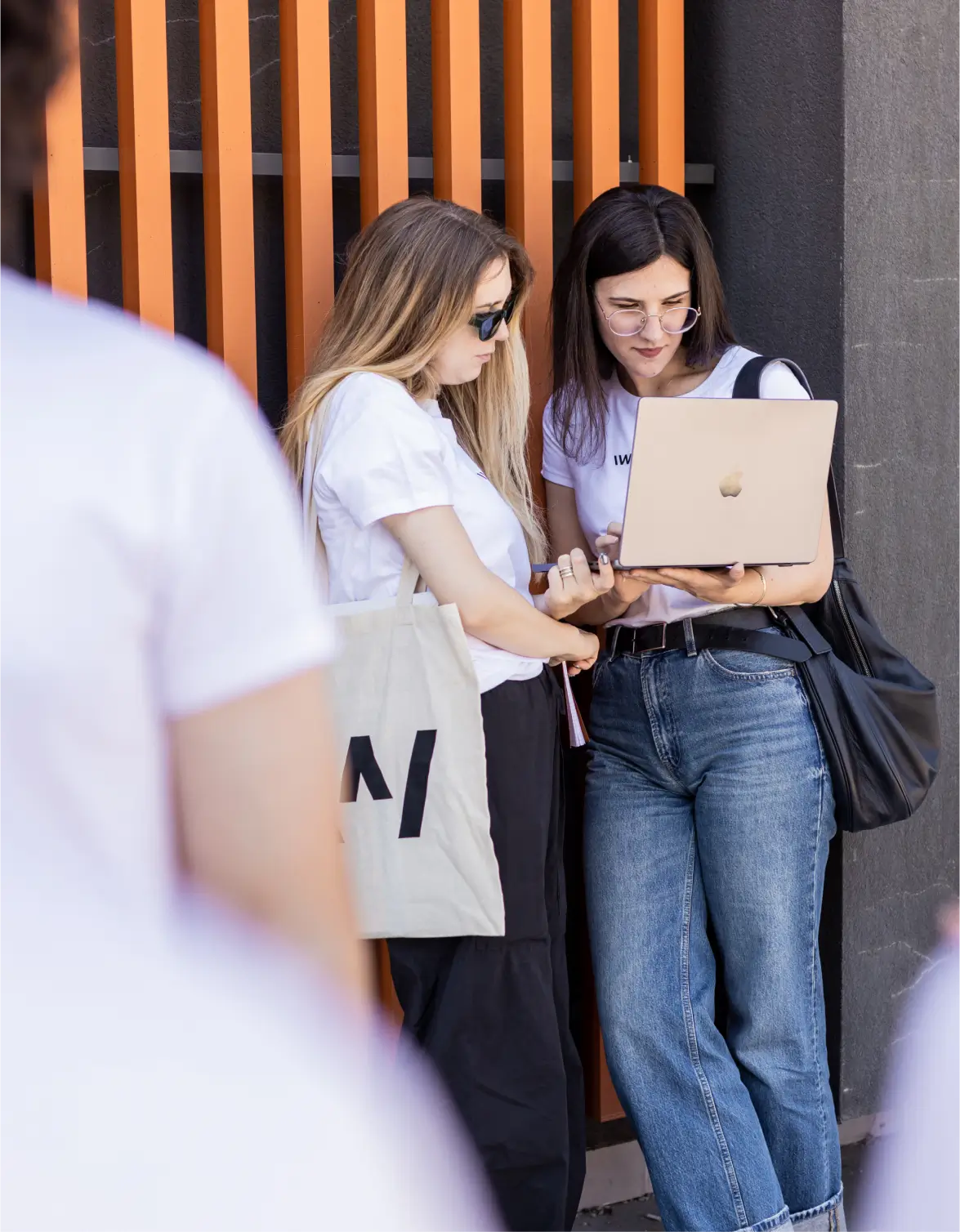 A candid, outdoor photograph focusing on two young women looking at a laptop. The woman on the right, wearing a white t-shirt, jeans, and glasses, holds a gold-colored laptop. The woman on the left, also in a white t-shirt, leans in to look at the screen while holding a large tote bag. They are standing next to a wall with orange vertical slat detailing. The foreground is blurred by the out-of-focus backs of two other people wearing white t-shirts.